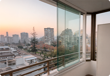 Evening city skyline viewed from a balcony with glass doors and a railing, trees and midrise buildings in the distance.
